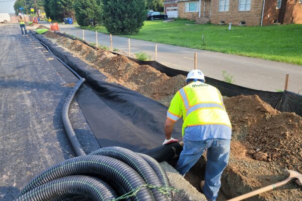 Workers laying a fabric layer down in a ditch for utilities outside of a neighborhood along a road.