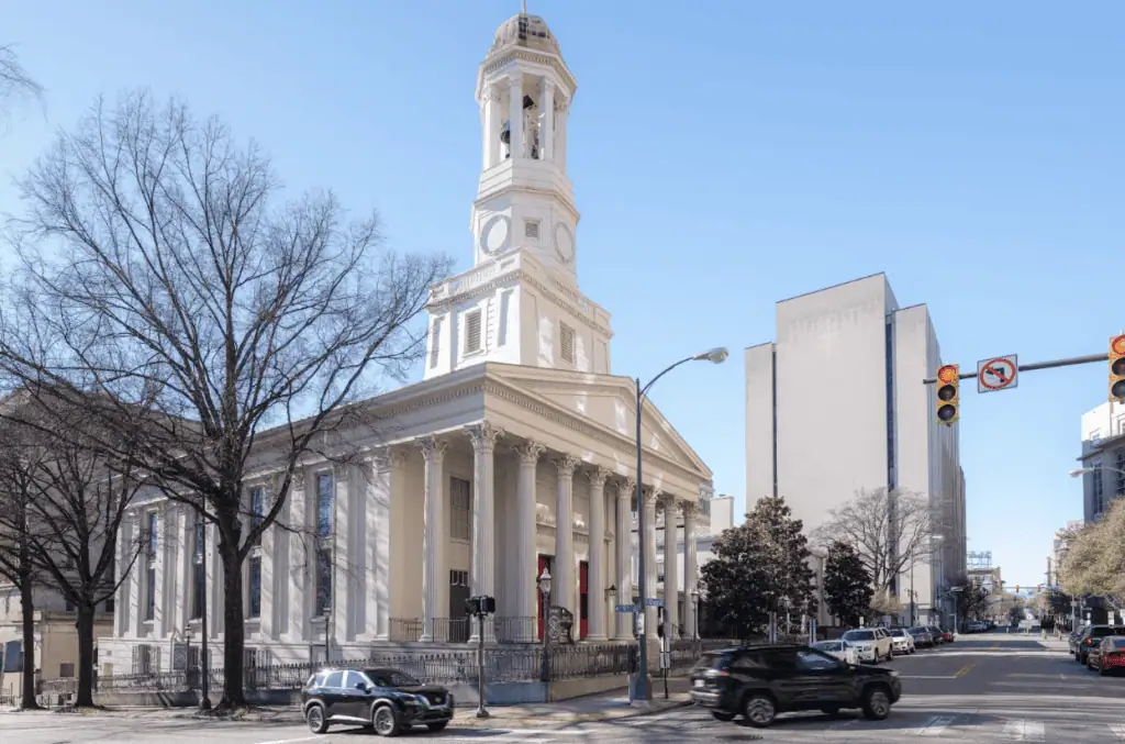 Historic white church with tall steeple and columned portico at a downtown street intersection, cars passing by under a clear blue sky.