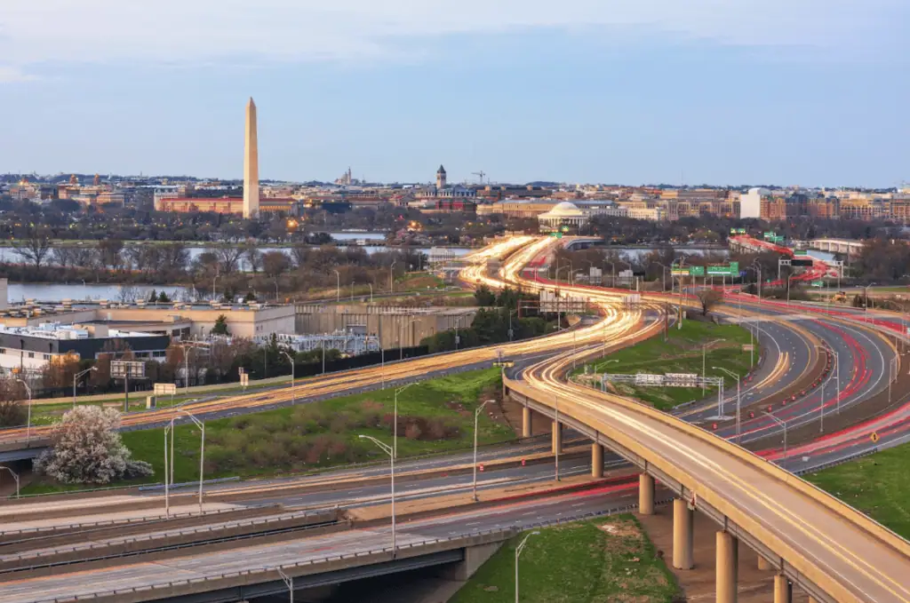 Aerial view of Washington, DC at dusk showing the Washington Monument and city skyline with curving highways below, captured with long-exposure light trails from moving traffic.