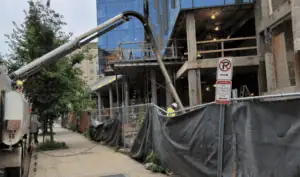 Concrete building under construction with a cement pump truck extending a hose into the structure, surrounded by fencing and sidewalk barriers in an urban area.