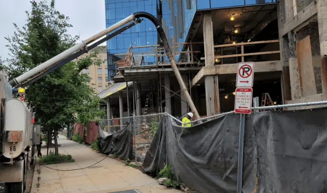 Concrete building under construction with a cement pump truck extending a hose into the structure, surrounded by fencing and sidewalk barriers in an urban area.