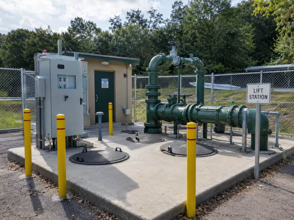Wastewater lift station with green discharge piping, access hatches, control equipment, and yellow safety bollards inside a fenced utility facility.